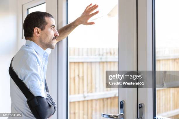 man wearing arm sling looking through window while standing at home - broken arm stock pictures, royalty-free photos & images