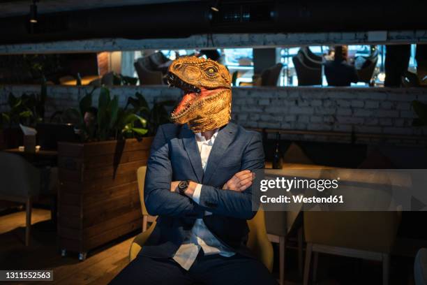 businessman wearing dinosaur mask while sitting with arms crossed in cafe - corporate business owner stock pictures, royalty-free photos & images