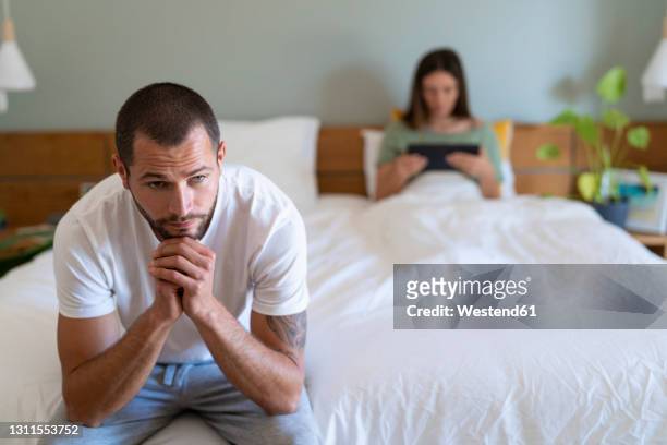 thoughtful man with hand on chin sitting on bed with girlfriend using digital tablet at home - man sadness stock pictures, royalty-free photos & images