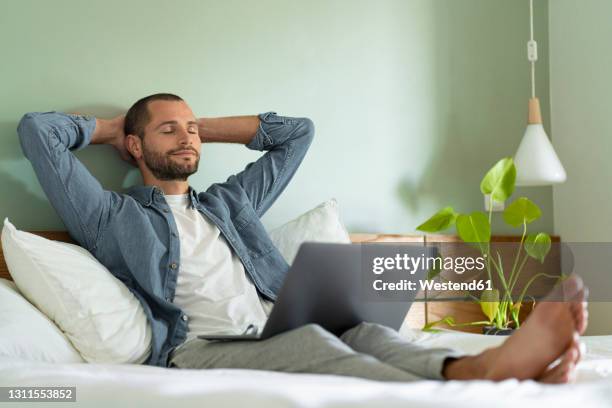 relaxed man sitting with laptop on lap in bedroom at home - hände hinter dem kopf stock-fotos und bilder