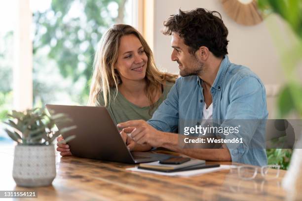 smiling couple looking at each other while sitting in front of laptop at table - couple photos et images de collection