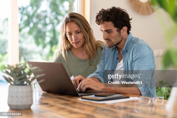 man using laptop while sitting by girlfriend at table - mid adult couple stock pictures, royalty-free photos & images