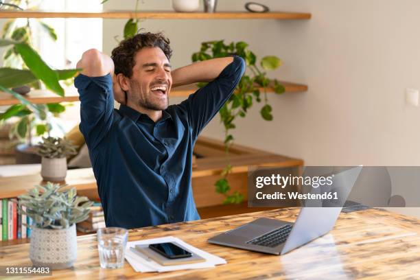 happy male freelancer with hands behind head laughing while sitting in front of laptop at home - person in front of computer stock pictures, royalty-free photos & images