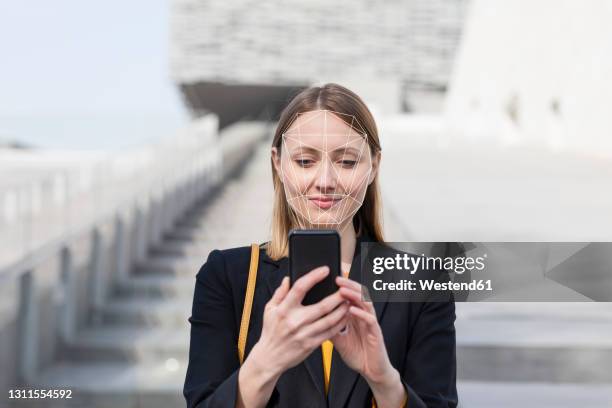 businesswoman unlocking mobile phone with face recognition technology while standing outdoors - biometrie stock-fotos und bilder