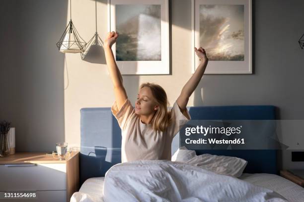 young woman stretching arms while sitting on bed at home - good morning stock pictures, royalty-free photos & images