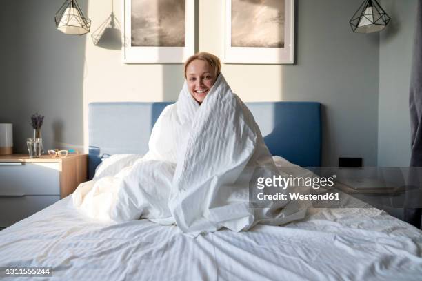 smiling woman wrapped in blanket sitting on bed at home - wrapped in a blanket stockfoto's en -beelden