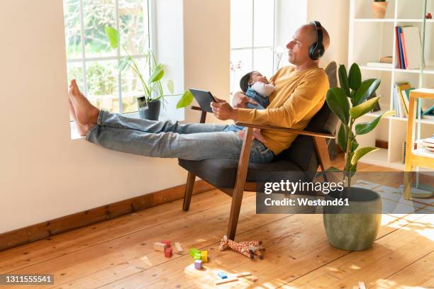 mid adult father looking away while listening music and holding sleeping baby at home - father holding sleeping baby imagens e fotografias de stock