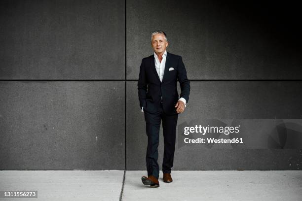 confident businessman with hand in pocket walking against gray wall - businessmann gehen im freien stock-fotos und bilder