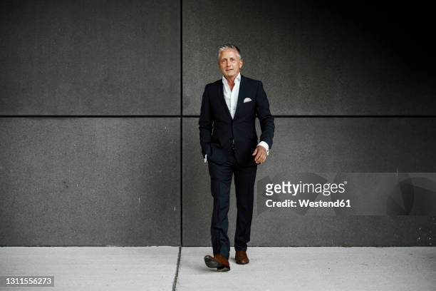 confident businessman with hand in pocket walking against gray wall - business men walking stock pictures, royalty-free photos & images