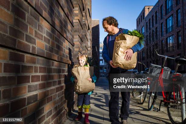 man with daughter holding paper bag of vegetables while walking on alley - shopping center stock-fotos und bilder