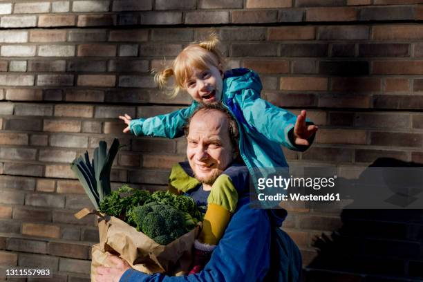 smiling man carrying cheerful daughter while holding bag of vegetables at wall - shopping paper bag stock-fotos und bilder