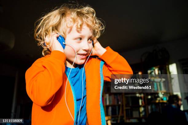 blond boy looking away while listening music through headphones at home - boy headphones stock-fotos und bilder