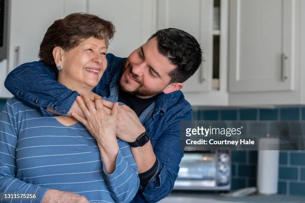 filho adulto abraça sua mãe mais velha enquanto ela cozinha uma refeição em casa na cozinha - filhos adultos - fotografias e filmes do acervo