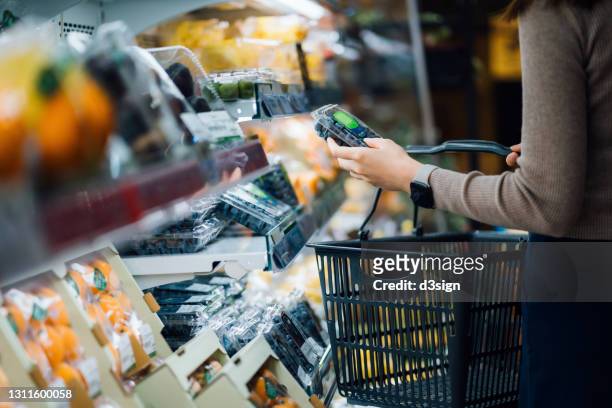 mid-section of young asian woman grocery shopping in a supermarket, carrying a shopping basket. she is choosing a pack of fresh organic blueberries in the produce aisle. healthy eating habits - blueberries fruit fotografías e imágenes de stock