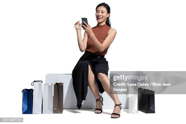 young woman using smart phone while sitting with shopping bags - chinese young adults shopping imagens e fotografias de stock