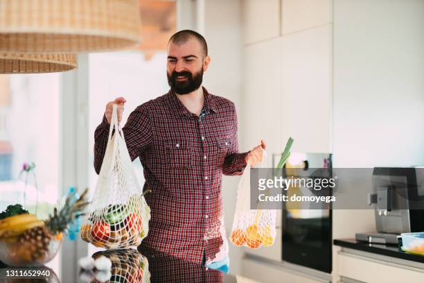 mens die herbruikbare boodschappenzak in de keuken thuis houdt - young man groceries kitchen stockfoto's en -beelden