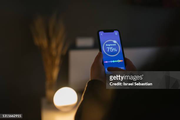 view from behind of a young woman using a smartphone app to adjust the lighting equipment in the bedroom of a modern home. - hand back lit stock pictures, royalty-free photos & images
