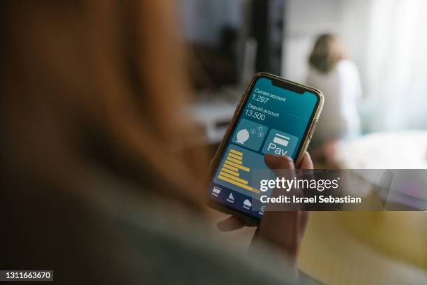 back view of an unrecognizable caucasian mother at home looking at her bank accounts from her cell phone.  while her daughter is playing in the living room. - online banking stock pictures, royalty-free photos & images