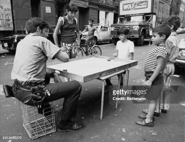 Patrolman William Mulligan playing 'Carom Hockey' with a young boy, both sitting on wire crates, as other children stand and watch on an unspecified...