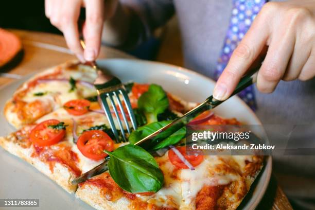 pizza margarita with mozzarella and basil leaves and pesto sauce, on a white plate, on the table in a cafe or fast food restaurant. nearby, a woman cuts off a slice of pizza to eat it. in the hands of cutlery. traditional italian cuisine. vegetarian food. - i was turning into a vegetable stock pictures, royalty-free photos & images