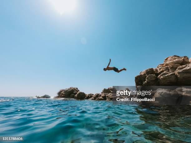millennial man is diving from a rock in the sea - italian beach fun stock pictures, royalty-free photos & images