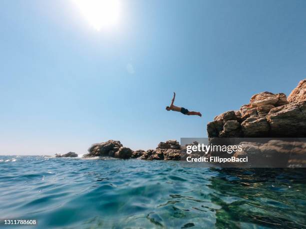 millennial man is diving from a rock in the sea - dove imagens e fotografias de stock