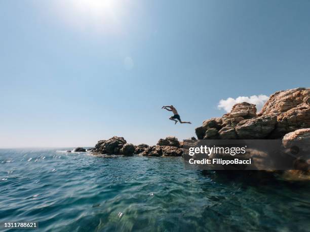 millennial man is diving from a rock in the sea - cannonball diving stock pictures, royalty-free photos & images
