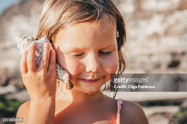 preschool girl with her eyes closed listens to sound of seashell on the beach - beach shells fotografías e imágenes de stock