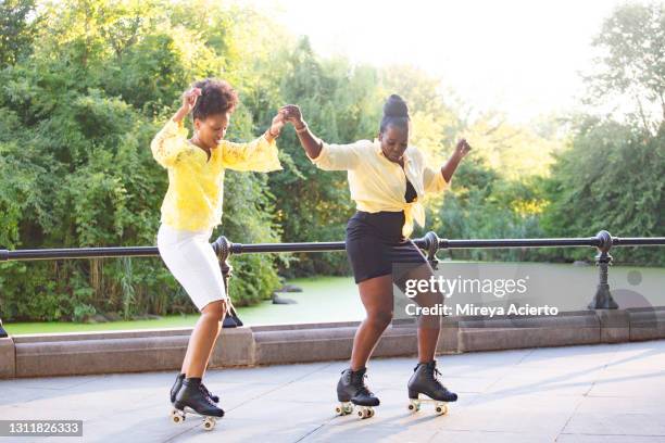 two african american senior women with their hair up in a bun, hold hands as they dance while roller skating in a public park near a pond, while wearing bright summer clothing. - summer hair bun stock pictures, royalty-free photos & images