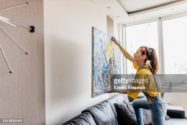 jovem mulher pendurando foto de arte na parede e sala de decoração - mulheres fotos - fotografias e filmes do acervo