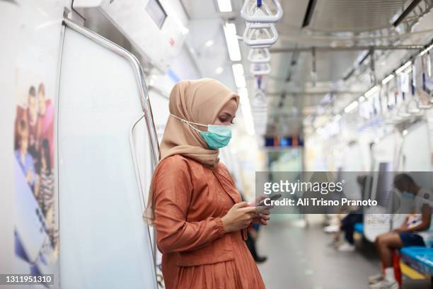 young asian hijab woman in mrt with protecting face mask using smartphone - indonesia covid stock pictures, royalty-free photos & images