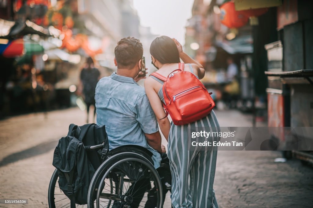 Rear view tourist asian chinese couple with wheelchair photographing using smart phone at petaling street, kuala lumpur during sunset