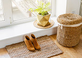 Hardwood floor with jute doormat, shoes and flower pot and seagrass laundry basket by window. Natural material objects in home concept. Home interior.