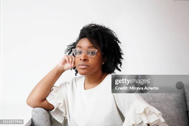 portrait of an african american millennial woman with curly hair, wears a white blouse and clear glasses,  while sitting on a grey couch in a brightly lit room. - grey sofa stock pictures, royalty-free photos & images