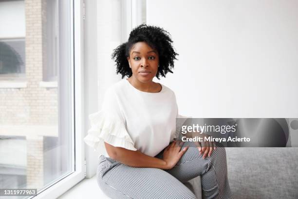 portrait of an african american millennial woman with curly hair, wears a white blouse while sitting on a grey couch in a brightly lit room. - brightly lit imagens e fotografias de stock