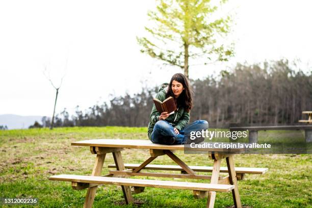 young woman sitting on top of a wooden table outdoors, reading a book. digital disconnection - mujer leyendo libro en el parque fotografías e imágenes de stock