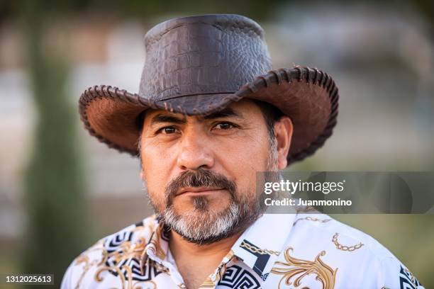 serious pensive mature hispanic man looking at the camera wearing a cowboy hat - mexican cowboy stock pictures, royalty-free photos & images