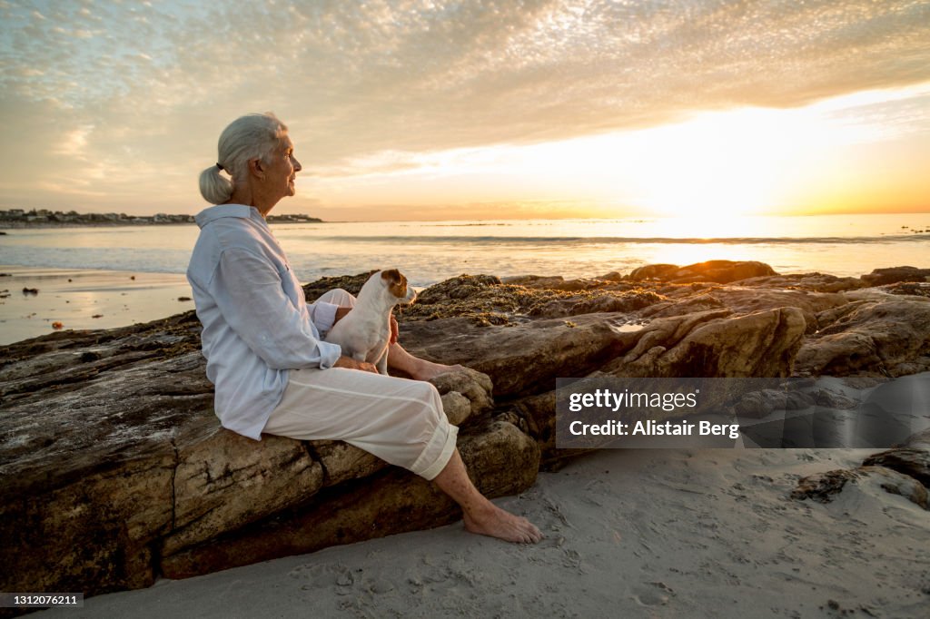 Senior woman sitting with her dog on a beach at sunset