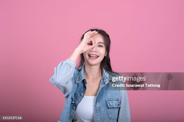 beautiful female smiling, demonstrating white teeth, looking at the camera through fingers in okay gesture. face expressions, emotions, and body language - okサイン　女性 ストックフォトと画像