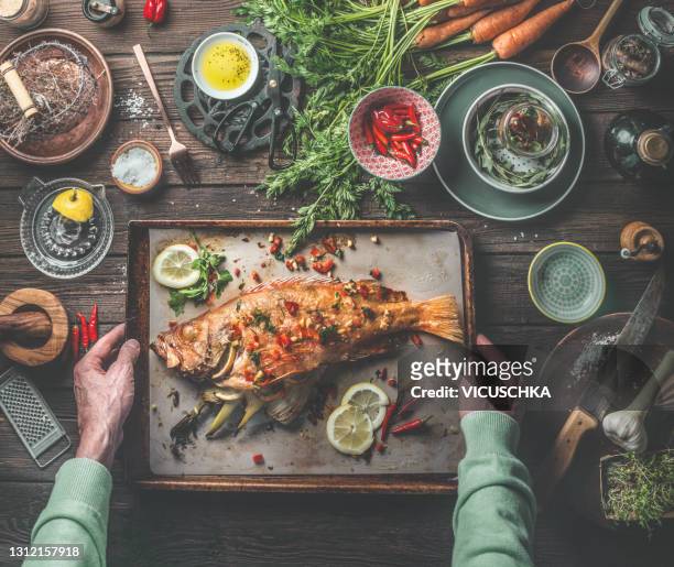 man's hands holding roasted whole redfish stuffed with fennel and lemon on baking tray on rustic table with fresh ingredients and kitchen utensils - mediterraanse gerechten stockfoto's en -beelden
