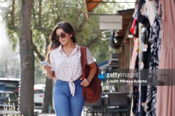 a latinx millennial woman with long brown hair and sunglasses, looks at her cell phone while walking through a small downtown shopping district while wearing a pink top with hearts, a backpack and jeans. - latin american and hispanic shopping bags bildbanksfoton och bilder