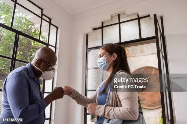 home caregiver arriving in patient's house and greeting with fist bump - wearing face mask - covid handshake stock pictures, royalty-free photos & images
