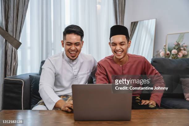 celebrating hari raya virtually. young men wearing malay traditional costume making online video call on laptop. - malay culture stock-fotos und bilder