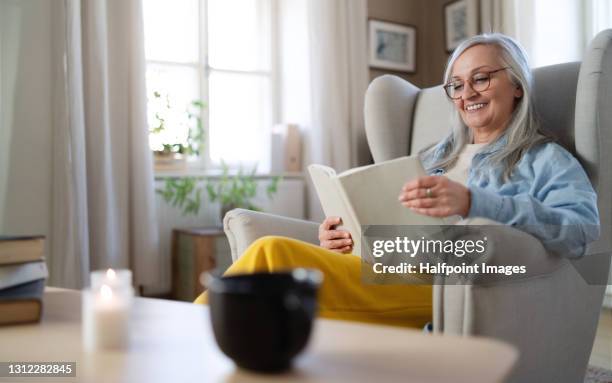 happy senior woman sitting on sofa at home, reading a book. - happy old women stockfoto's en -beelden