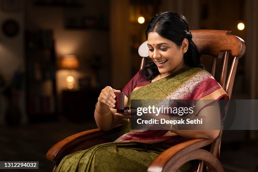 shot of a woman in sari got shocked while opening the diamond box of ring sitting on a rocking chair at home:- stock photo