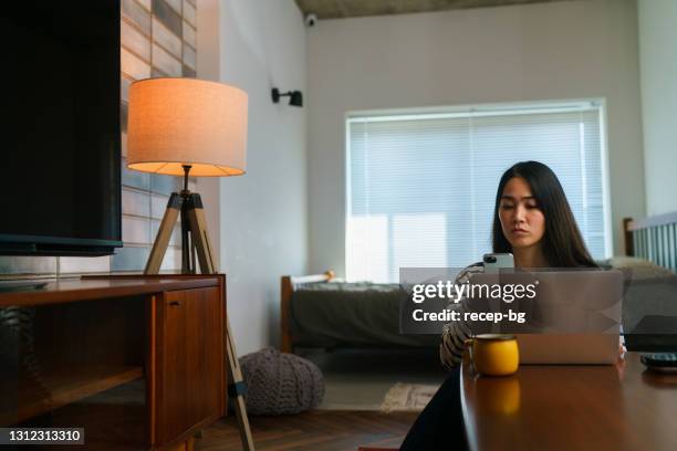 young woman using her smart phone app to turn off the light while working on laptop in living room at home - bluetooth stock pictures, royalty-free photos & images