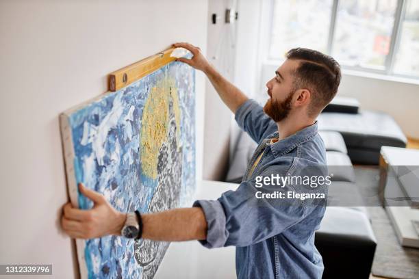 man with beard hanging a painting on the wall at his living room - hanging stock pictures, royalty-free photos & images