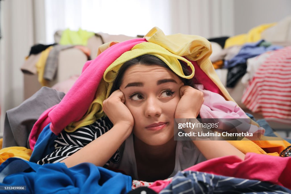 Pensive young woman with lots of clothes on floor in room. Fast fashion