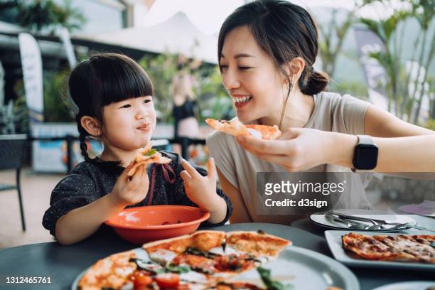 joyful young asian mother and lovely little daughter enjoying pizza lunch in an outdoor restaurant. family enjoying bonding time and a happy meal together. family and eating out lifestyle - family cafe foto e immagini stock