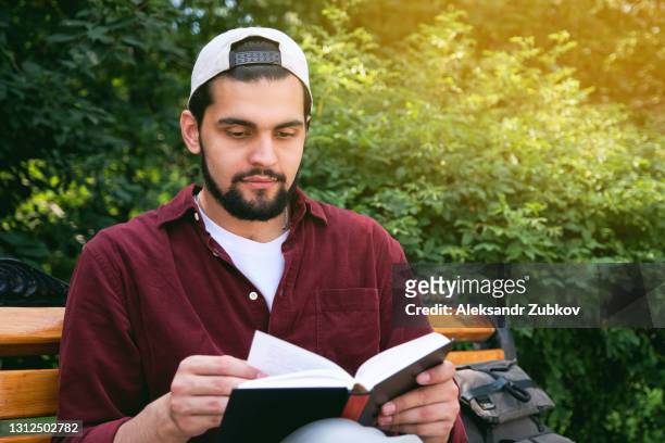 a young indian man sits on a bench in a public park with a book in his hands, on a sunny summer day against a background of green trees. a dark-skinned indian student in a cap reads, prepares for the exam and admission to an international university. - international student day stock-fotos und bilder